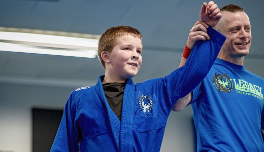 Young boy in blue martial arts gi celebrating victory at Legacy Jiu-Jitsu Academy.